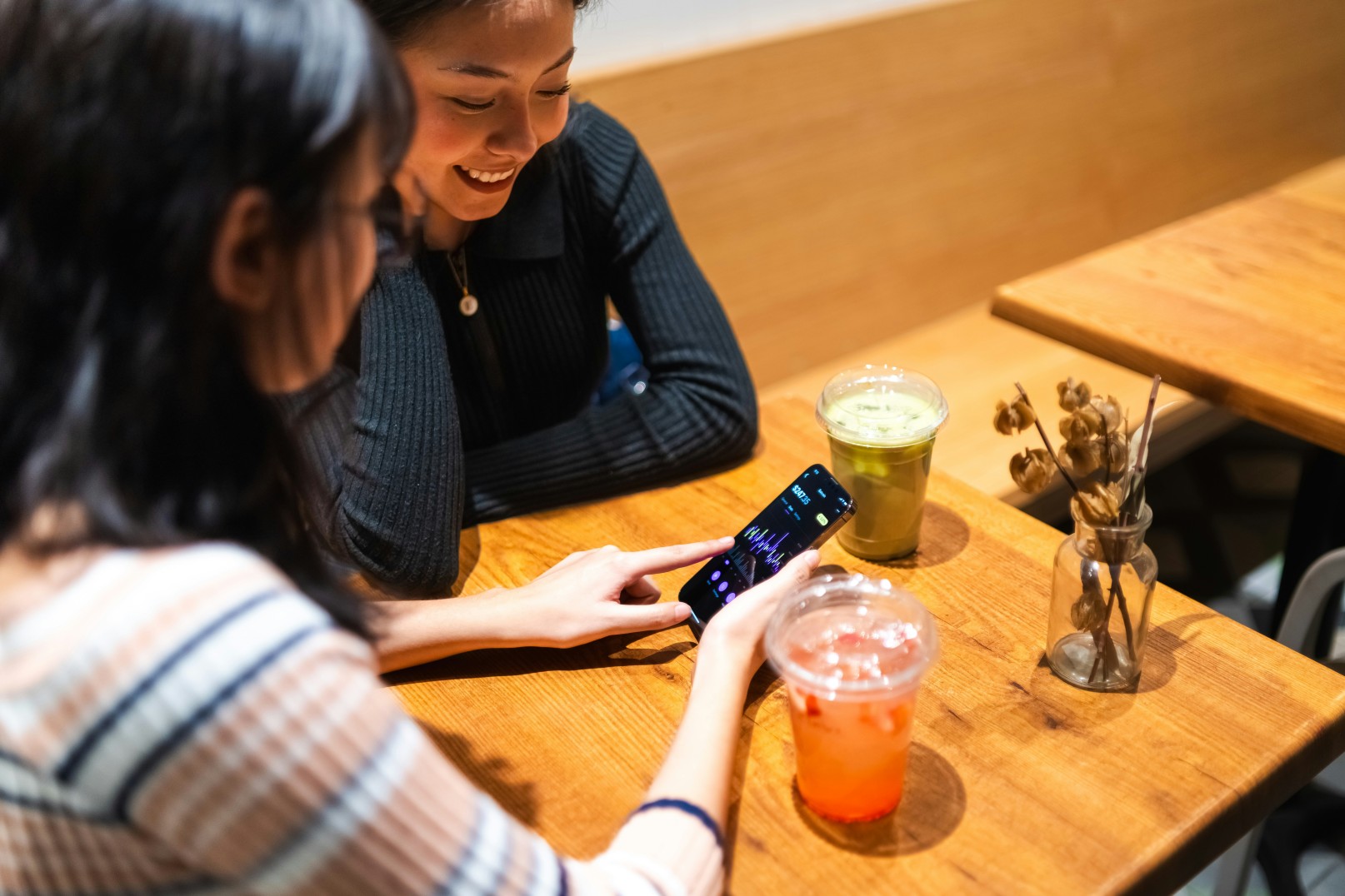 Duas pessoas conversando em uma cafeteria enquanto uma delas mostra algo no celular, sobre uma mesa com bebidas.